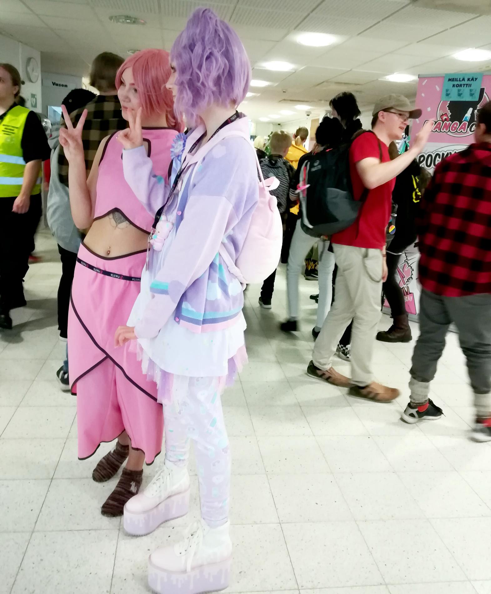 Kaksi nuorta poseeraamassa cosplay-asuissa toiselle kameralle tapahtumassa. Ihmisiä käytävällä.  