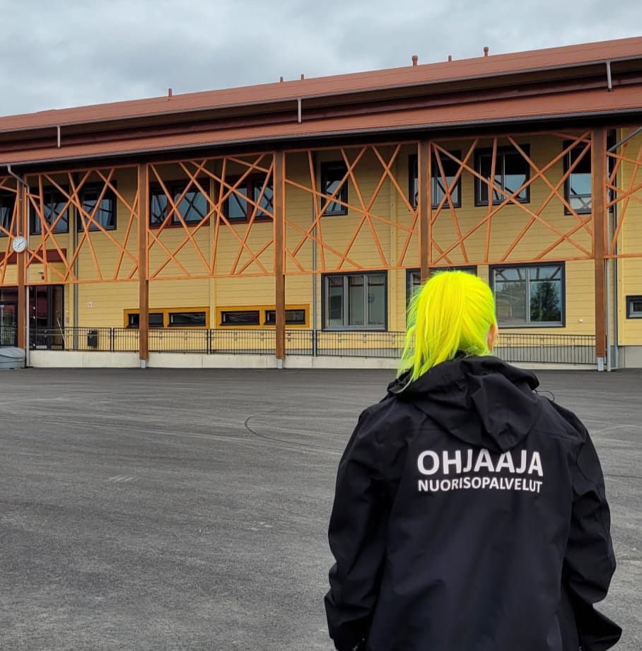 Keltahiuksinen nainen kävelemäsää musta ohjaaja takki päällä kohti rakennusta.