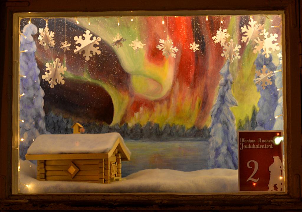 Jouluinen asetelma, taustalla revontulimaalaus ja etualalla lumikattoinen mökki sekä lumihiutaleita.