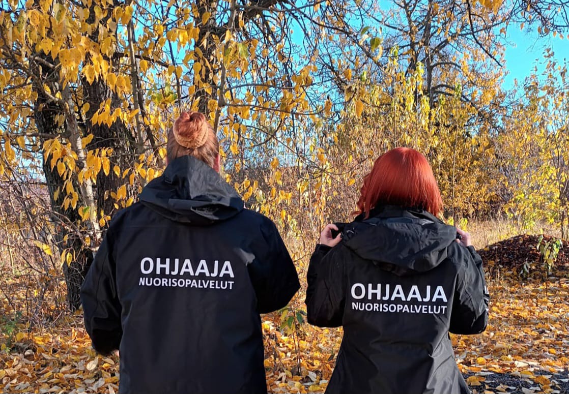 Kaksi henkilöä seisoo vierekkäin selkä kuvaajaa kohti. Heillä on päällään samanlaiset takit, joissa lukee teksti: Ohjaaja, nuorisopalvelut.