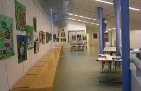 Näyttelytila ylätasanteella, vasemmalla seinällä tauluja ja pitkä puinen pöytä, oikealla pylväitä.